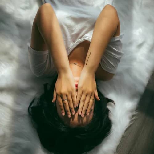 woman with black hair and wearing a white shirt with hands over her eyes lying in bed. Image denotes that she is distressed about sleeping and wondering what is insomnia